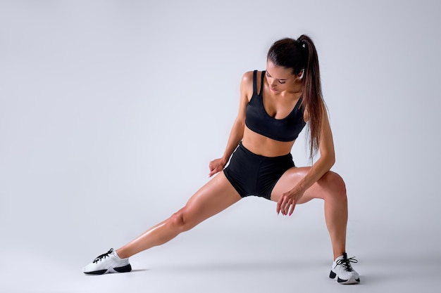 Retrato de una joven morena estirando las piernas antes del entrenamiento, aislada en gris