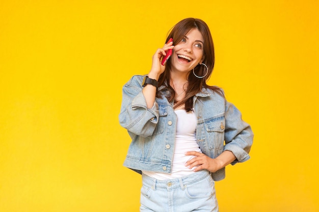 Retrato de una joven morena feliz y emocionada con un estilo informal de mezclilla de pie, con la mano en la cintura, mirando hacia otro lado y hablando por su teléfono móvil. tiro de estudio interior, aislado sobre fondo amarillo.