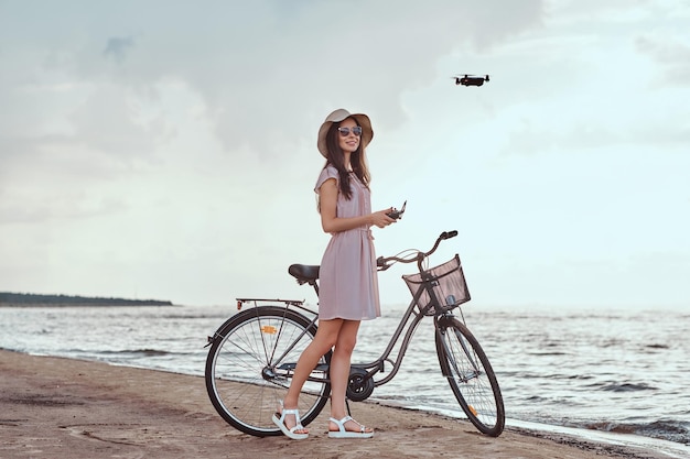Retrato de una joven morena con gafas de sol y sombrero vestido que disfruta de las vacaciones en la playa, jugando con el cuadricóptero.