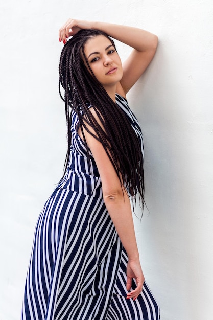 Retrato de una joven morena seria con peinado de rastas afroamericanas negras con vestido a rayas posando y mirando a la cámara. foto de estudio interior en la pared blanca.