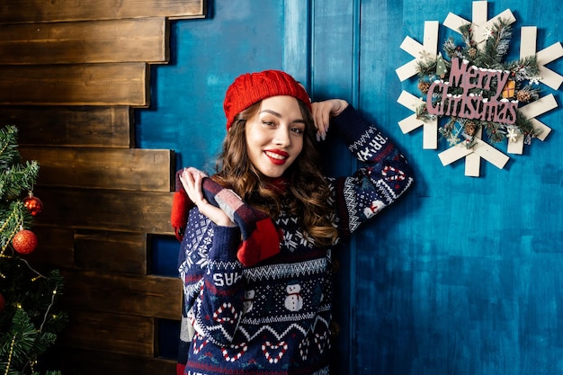 Retrato de una joven morena sonriendo en una bufanda de suéter de sombrero de Año Nuevo contra el fondo de una pared azul de madera La Feliz Navidad