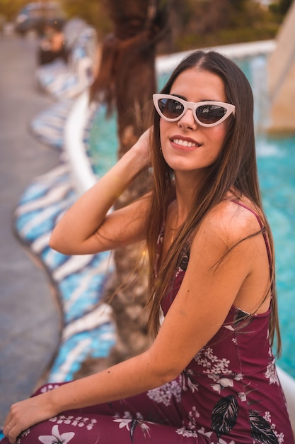 Retrato de una joven morena con un vestido floral marrón disfrutando del verano en la hora dorada por una fuente de la ciudad