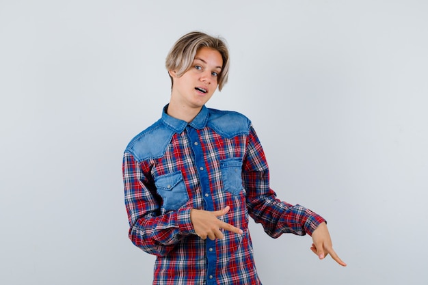 Foto retrato de joven muchacho adolescente apuntando hacia abajo en camisa a cuadros y mirando curiosa vista frontal
