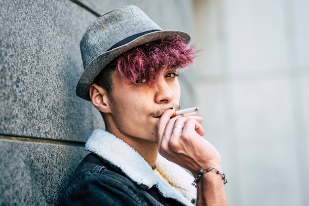 Retrato de joven muchacho adolescente fumando tabaco - estilo de vida poco saludable y gente masculina de moda - cabello de estilo alternativo de diversidad violeta - mirando a cámara - fondo urbano