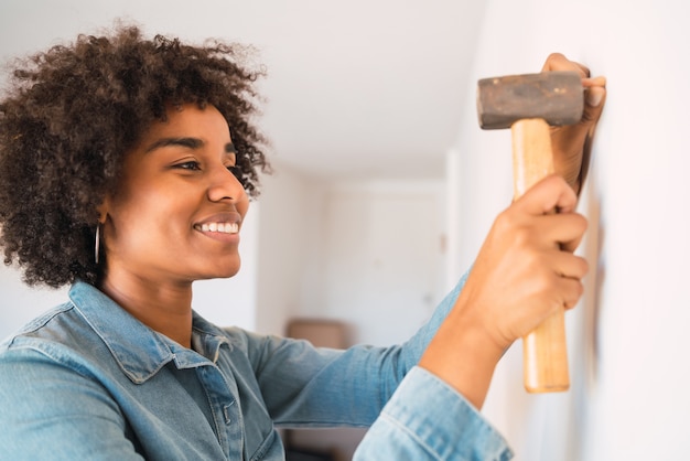 Retrato de joven mujer afro martillando clavos en la pared en casa