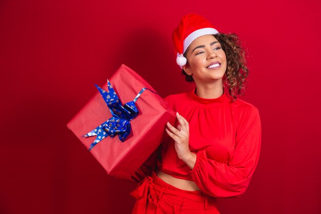 Retrato de joven mujer afro con sombrero de santa claus sosteniendo un regalo sobre fondo rojo. concepto de noche de navidad