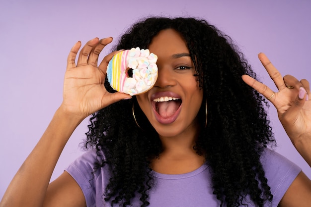 Retrato de joven mujer afro sosteniendo una rosquilla y divirtiéndose mientras está de pie sobre un fondo aislado. Concepto de dulces y alimentos.