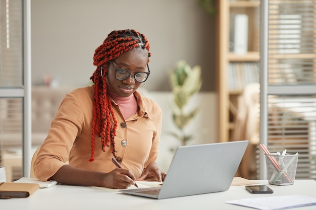 Retrato de joven mujer afroamericana escribiendo en el planificador mientras trabaja o estudia en el escritorio en la oficina en casa, espacio de copia