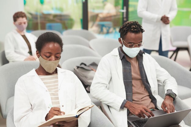 Retrato de joven mujer afroamericana con máscara y bata de laboratorio mientras escucha una conferencia sobre medicina en la universidad o centro de coworking, espacio de copia