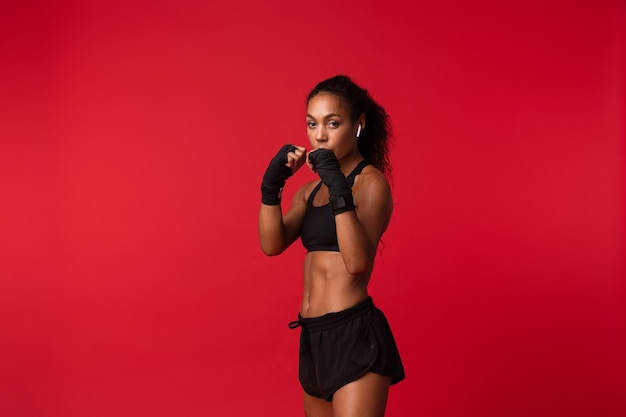 Retrato de joven mujer afroamericana en ropa deportiva negra de boxeo con vendas deportivas en sus manos, aislado sobre pared roja