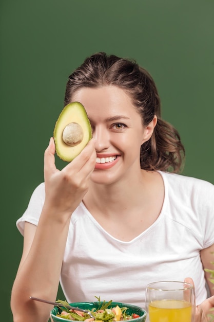 Retrato de una joven mujer alegre con aguacate en la mesa llena de vegetales crudos saludables y frutas...