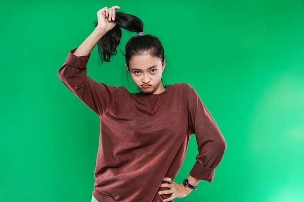 Retrato joven mujer asiática expresando el ceño fruncido mientras sostiene su largo cabello negro y mirando a la cámara en la pared verde