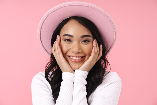 Retrato de joven mujer asiática morena con sombrero sonriendo y mirando a cámara aislada sobre pared rosa