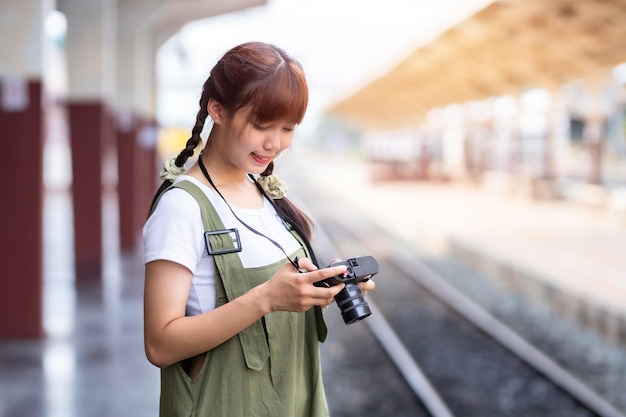 Retrato Joven mujer asiática sonriente turista Chica viajera caminando y sosteniendo la cámara espera el viaje en tren se toma en la plataforma ferroviaria Tailandia verano relax vacaciones Concepto