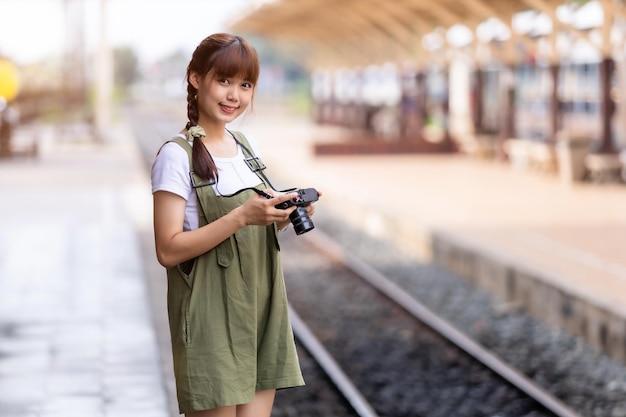 Retrato Joven mujer asiática sonriente turista Chica viajera caminando y sosteniendo la cámara espera el viaje en tren se toma en la plataforma ferroviaria Tailandia verano relax vacaciones Concepto