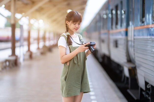 Retrato Joven mujer asiática sonriente turista Chica viajera caminando y sosteniendo la cámara espera el viaje en tren se toma en la plataforma ferroviaria Tailandia verano relax vacaciones Concepto