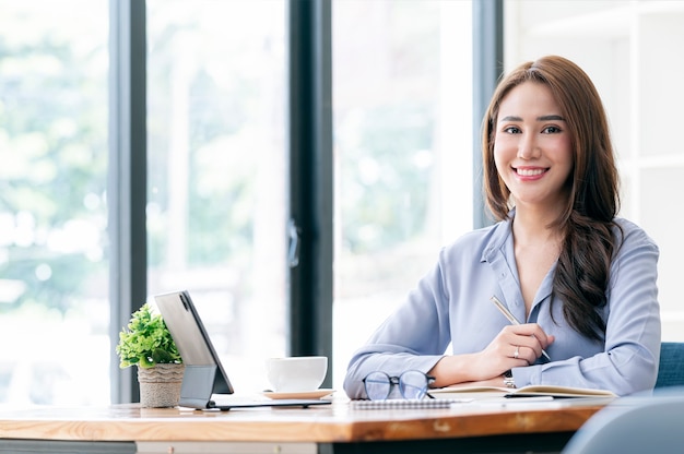 Retrato de joven mujer asiática sosteniendo la pluma, sonriendo y mirando a la cámara mientras está sentada en la oficina de su escritorio.