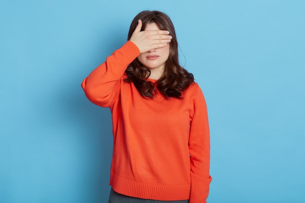 Retrato de joven mujer asustada bastante caucásica que cubre los ojos con las manos mientras está de pie contra el azul