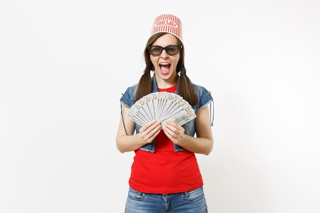 Retrato de joven mujer atractiva llena de alegría en gafas 3d con cubo para palomitas de maíz en la cabeza viendo una película, sosteniendo un paquete de dólares, dinero en efectivo aislado sobre fondo blanco. Emociones en el cine.