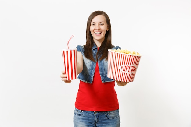 Retrato de joven mujer atractiva sonriente en ropa casual viendo una película, mostrando un cubo de palomitas de maíz y un vaso de plástico de refresco o cola en la cámara aislada sobre fondo blanco. Emociones en el cine.