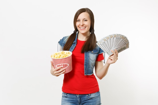 Retrato de joven mujer atractiva sonriente en ropa casual viendo una película, sosteniendo un cubo de palomitas de maíz y un paquete de dólares, dinero en efectivo aislado sobre fondo blanco. Emociones en el concepto de cine.