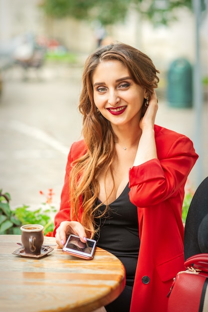 Retrato de una joven mujer blanca sentada a la mesa con un teléfono inteligente y mirando a la cámara al aire libre.