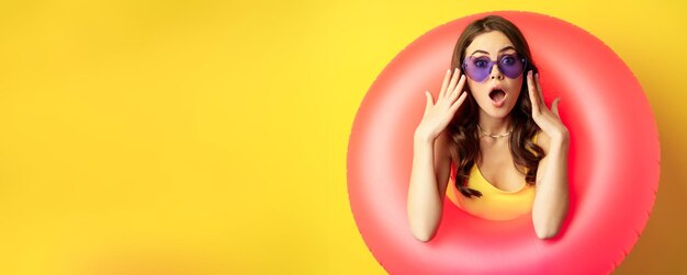 Retrato de una joven mujer bonita con gafas de sol usando un anillo de natación en la playa que parece sorprendida y asombrada