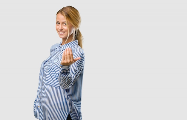 Retrato de una joven mujer bonita rubia que invita a venir, confiada y sonriente haciendo un gesto con la mano