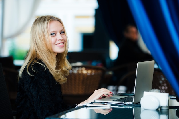 Retrato de joven mujer bonita rubia sentada en la calle café con su computadora portátil, escribiendo y sonriendo