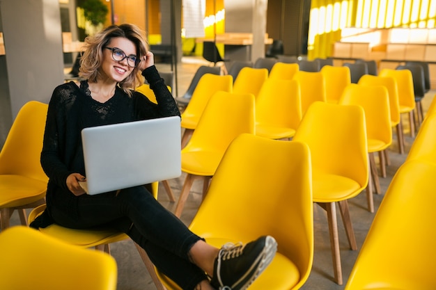 Retrato de joven mujer bonita sentada en la sala de conferencias, trabajando en equipo portátil, con gafas