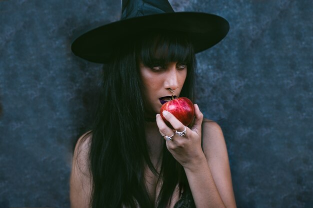 Foto retrato joven de la mujer de la bruja de halloween que come una manzana roja. belleza vampiro enojado bruja dama con boca negra en la oscuridad, con un sombrero de bruja.