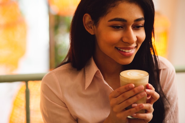 Retrato de una joven mujer de cabello oscuro complacida que se siente bien y sonriendo mientras tiene un vaso de café con leche en sus manos