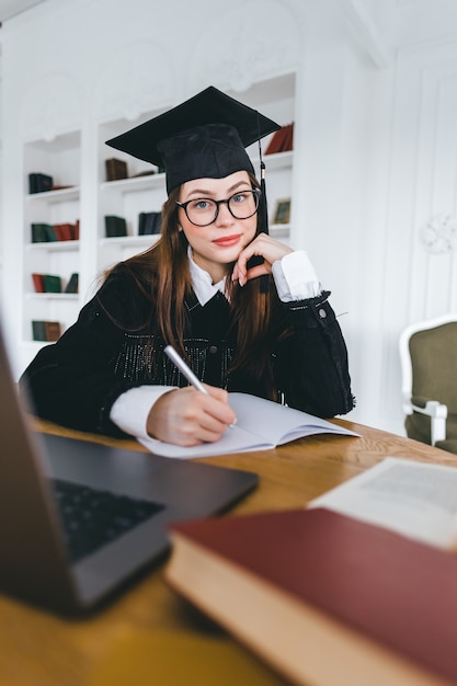 Retrato de joven mujer caucásica en anteojos estudiante universitario en traje de graduación tomando notas