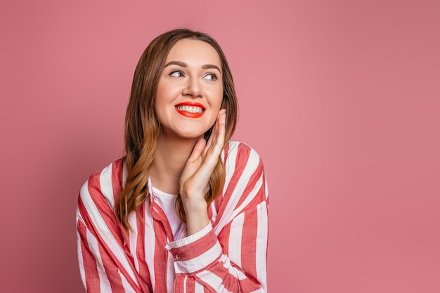 Retrato de joven mujer caucásica de cabello castaño con labios rojos con camisa roja a rayas sonriendo y mirando hacia el lado aislado en la pared rosa.