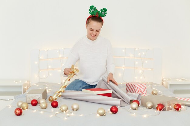 Retrato de joven mujer caucásica feliz y dulce con jersey de estilo casual y jeans, sentada en la cama preparando regalos de Navidad envolviendo cajas de regalo y agregando cintas.