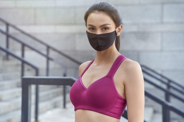 Retrato de una joven mujer caucásica de fitness con mascarilla protectora mirando a la cámara mientras corre al aire libre durante la pandemia de coronavirus. Covid 19 y actividad física, deporte, estilo de vida saludable