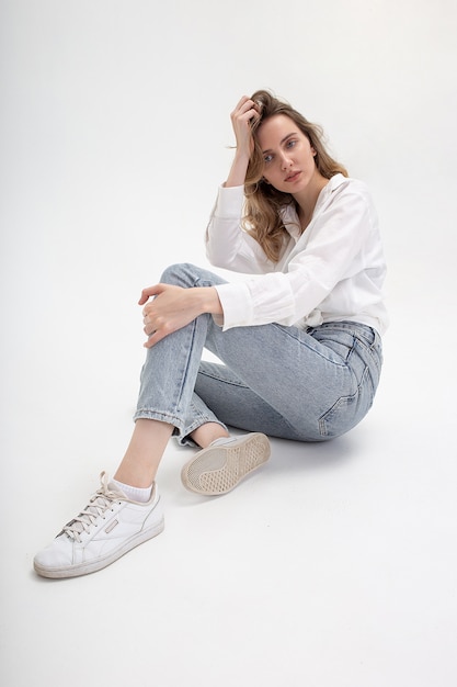 Retrato de joven mujer caucásica pensativa posando en camisa y jeans, sentado en blanco