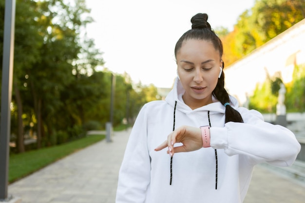 Retrato de una joven mujer delgada en una sudadera con capucha blanca escucha música con auriculares y mira el reloj. Entrenamiento matutino al aire libre