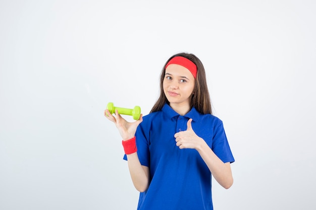 Retrato de joven mujer deportiva en camiseta azul sosteniendo pesas y dando pulgar hacia arriba.