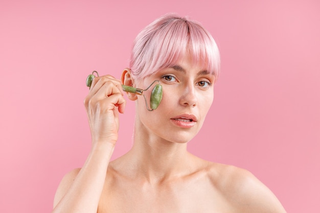 Retrato de joven mujer desnuda con cabello rosado con rodillo facial de jade natural para el cuidado de la piel posando