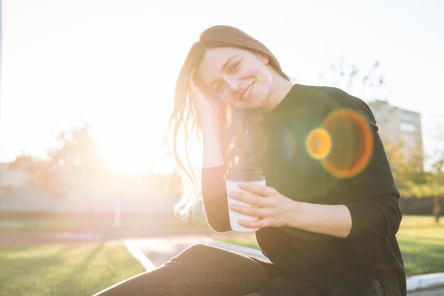 Retrato de una joven mujer feliz con el pelo largo con una taza de café de papel en el parque de la ciudad en la hora dorada