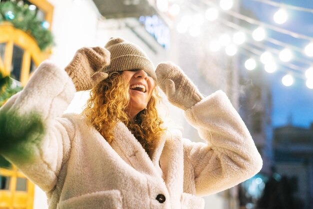 Retrato de una joven mujer feliz con el pelo rizado en una chaqueta de piel divirtiéndose en la calle de invierno decorada con luces