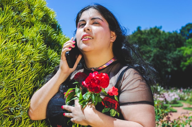Retrato de joven mujer latina hispana caucásica enamorada dramática en un parque público con rosas y árboles ella está de pie hablando por teléfono abrazando algunas flores