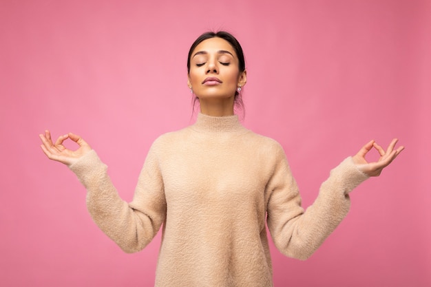 Retrato de joven mujer morena atractiva feliz tranquila positiva con emociones sinceras vistiendo jersey beige aislado sobre fondo rosa con espacio de copia y haciendo mediación. Concepto de yoga.