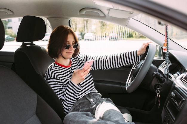 Foto retrato de una joven mujer morena conduciendo en un coche usando un teléfono inteligente