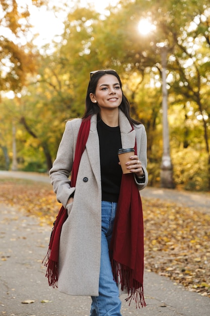 Retrato de joven mujer morena vistiendo abrigo bebiendo café para llevar y sonriendo mientras camina en el parque de otoño