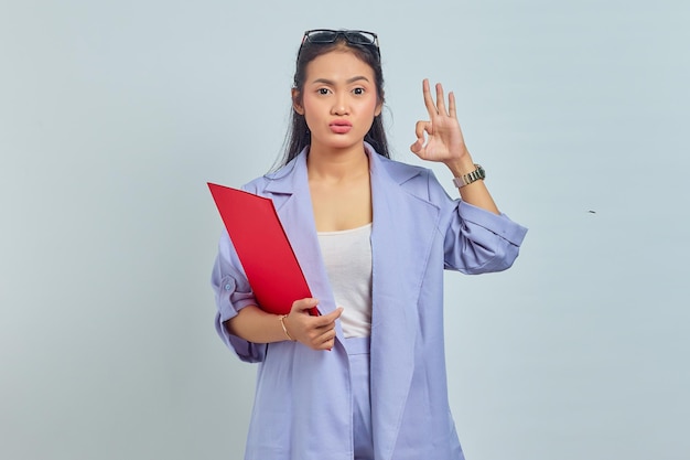 Retrato de una joven mujer de negocios asiática positiva con traje que sostiene una carpeta de documentos y hace un gesto correcto, está de acuerdo con una buena idea aislada en un fondo morado