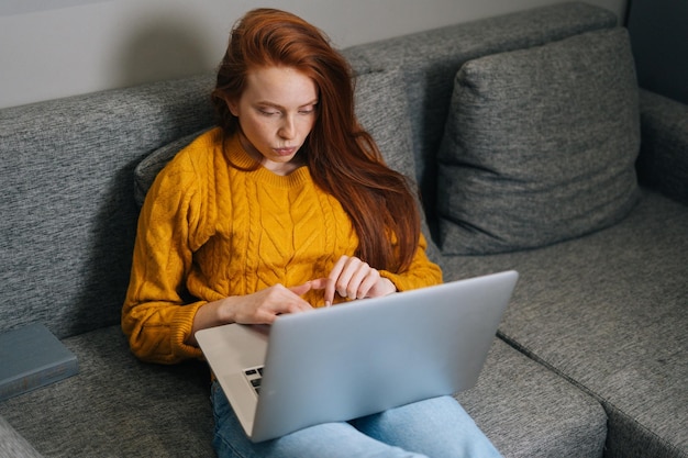 Retrato de una joven mujer de negocios cansada escribiendo en una computadora portátil sentada en un sofá en la sala de estar