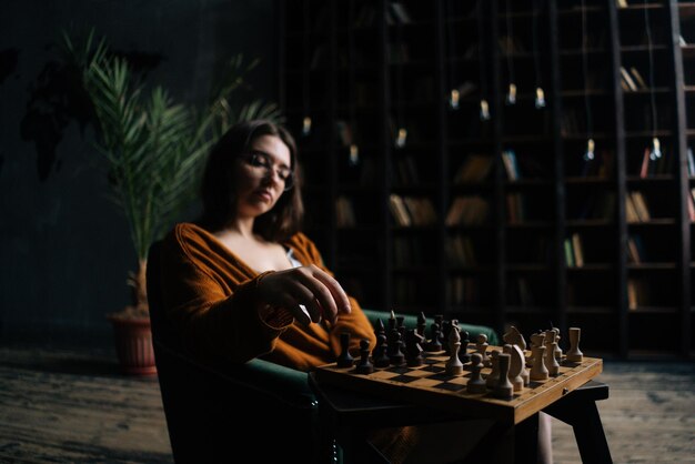 Retrato de una joven mujer de negocios confiada que usa anteojos elegantes haciendo que el ajedrez se mueva sentado en un sillón en la oscura sala de la biblioteca