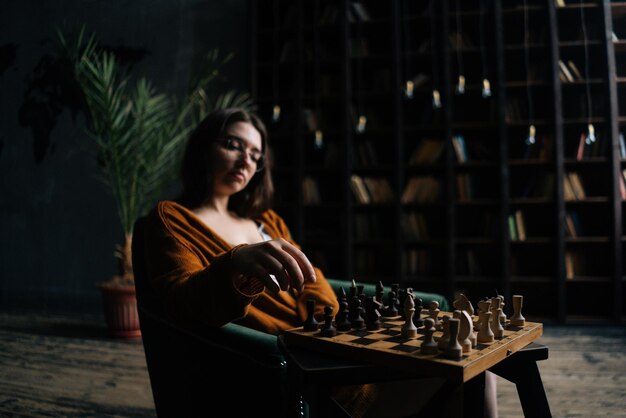 Retrato de una joven mujer de negocios confiada que usa anteojos elegantes haciendo que el ajedrez se mueva sentado en un sillón en la oscura sala de la biblioteca